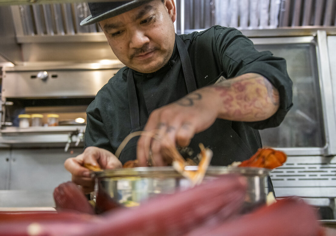 Cambodian chef known as "Chef T" wears his black chef uniform and a fedora while preparing a lobster tail in a restaurant kitchen.