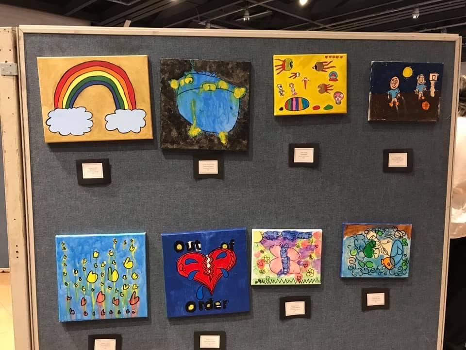 Childlike art hangs from a wall. The art features painted images such as a rainbow, flowers and a butterfly.