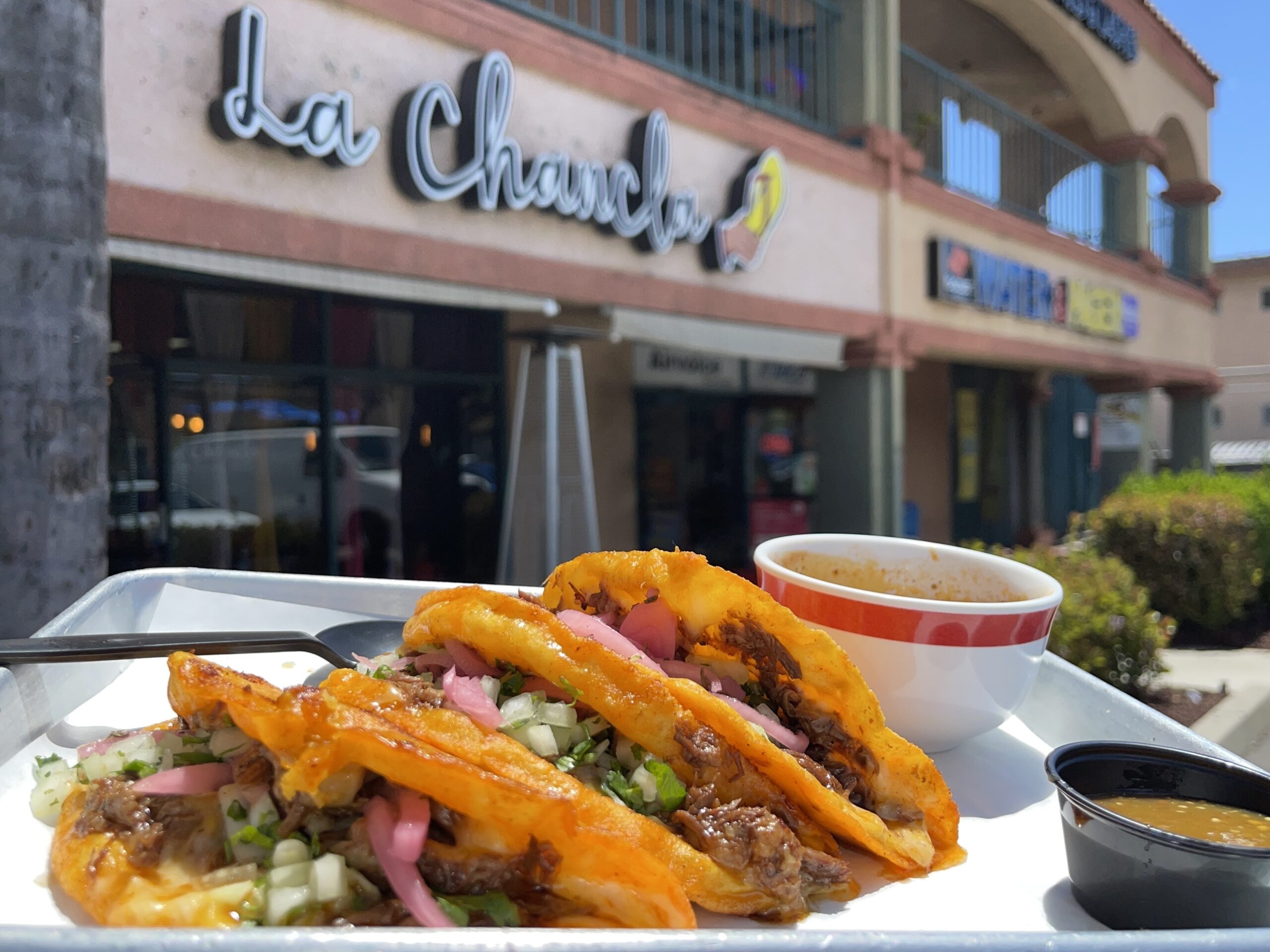 A plate of tacos is seen on a table outside a building that says "La Chancla."