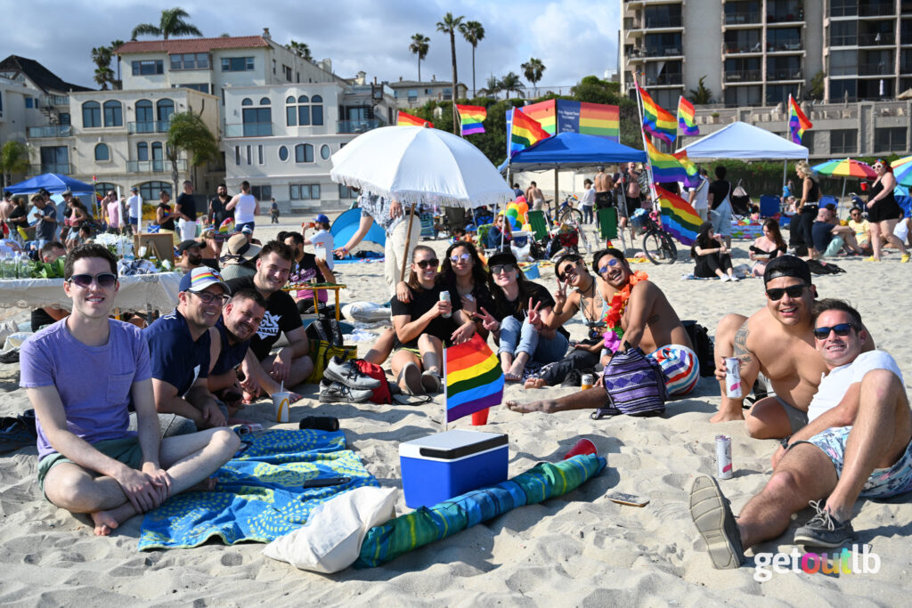Dozens of people sit on the beach that is decorated with rainbow flags.