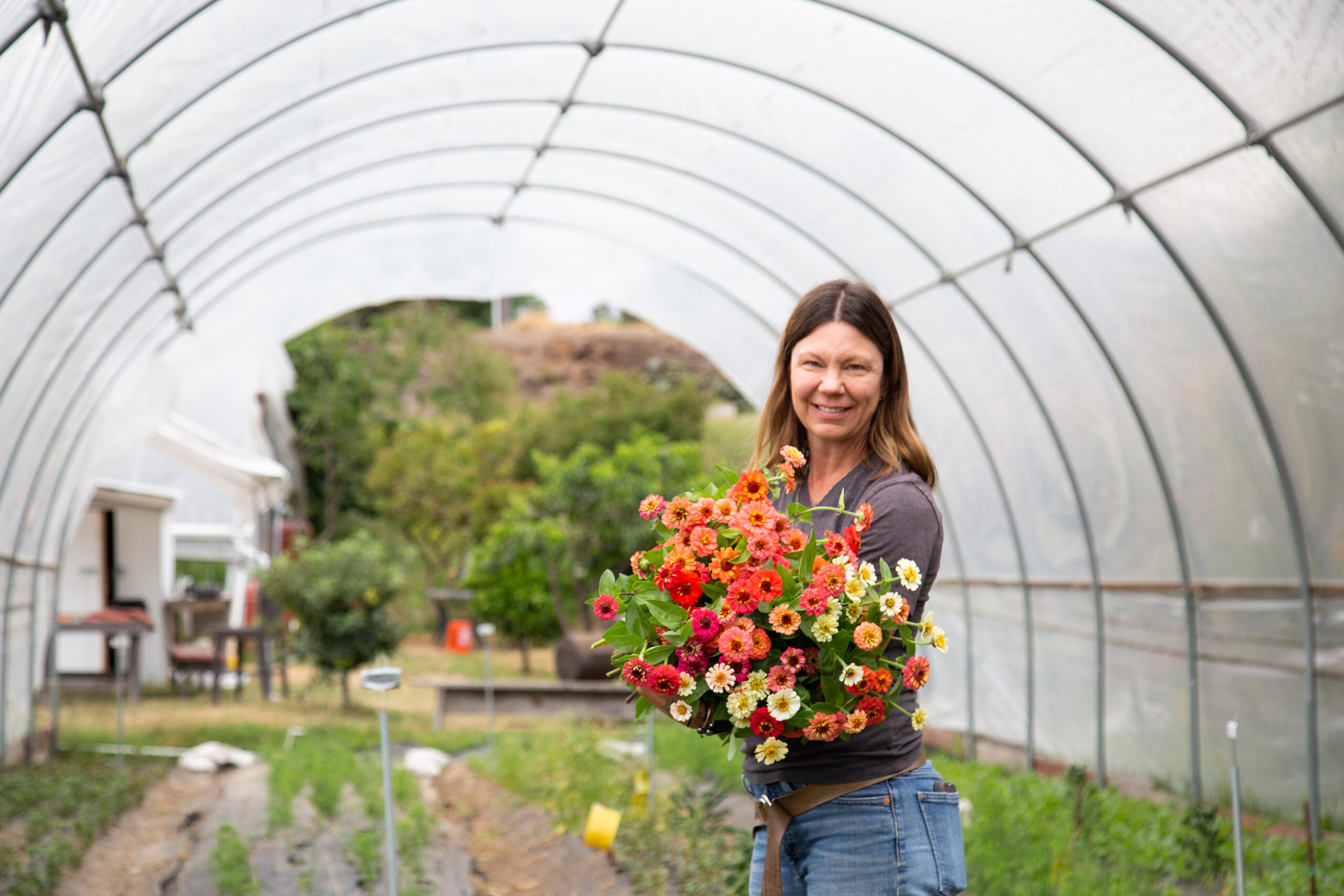 A woman wearing jeans and a dark T-shirt smiles while holding a bouquet of red, pink, yellow, white, and orange flowers. A farm is blurred behind her.
