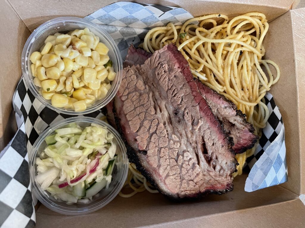Two pieces of fatty brisket on top of garlic noodles with two containers of corn and pickled, shredded cabbage and cucumber.