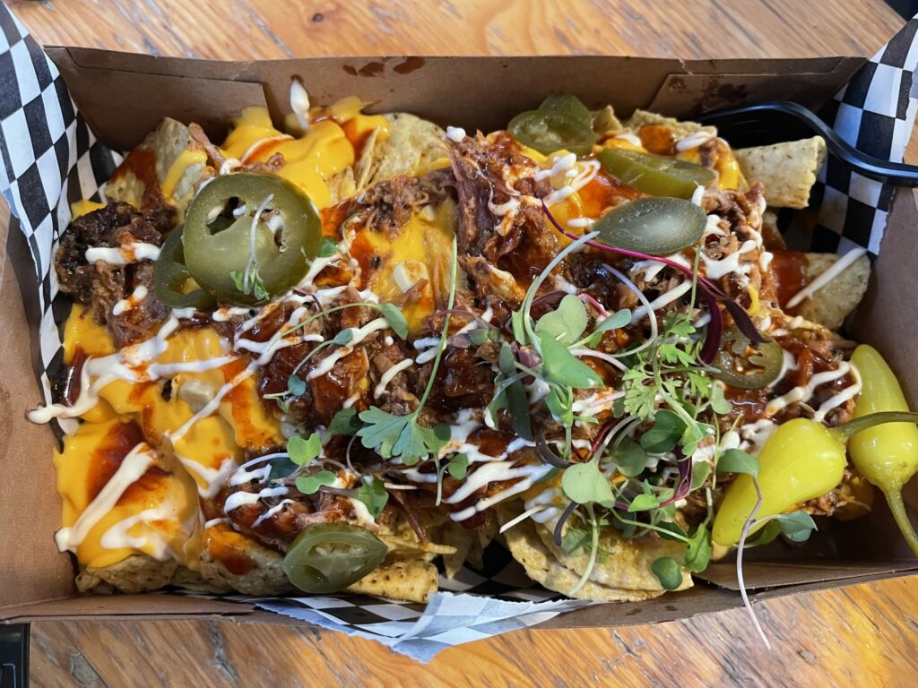 Nachos in take out container with melted cheese, shredded meat with barbecue sauce then topped with jalapeños, crema, and micro greens.