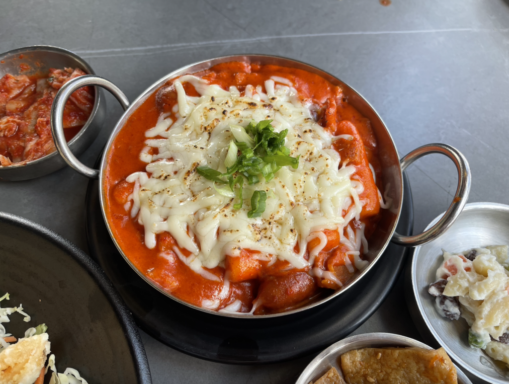 Rose tteokbokki is served in a metal wide bowl with handles. Cylinder strips of rice cakes and mini sausages are covered in a light red sauce. Mozzarella cheese is melted on top and slightly charred. The dish is topped with sliced green onion.