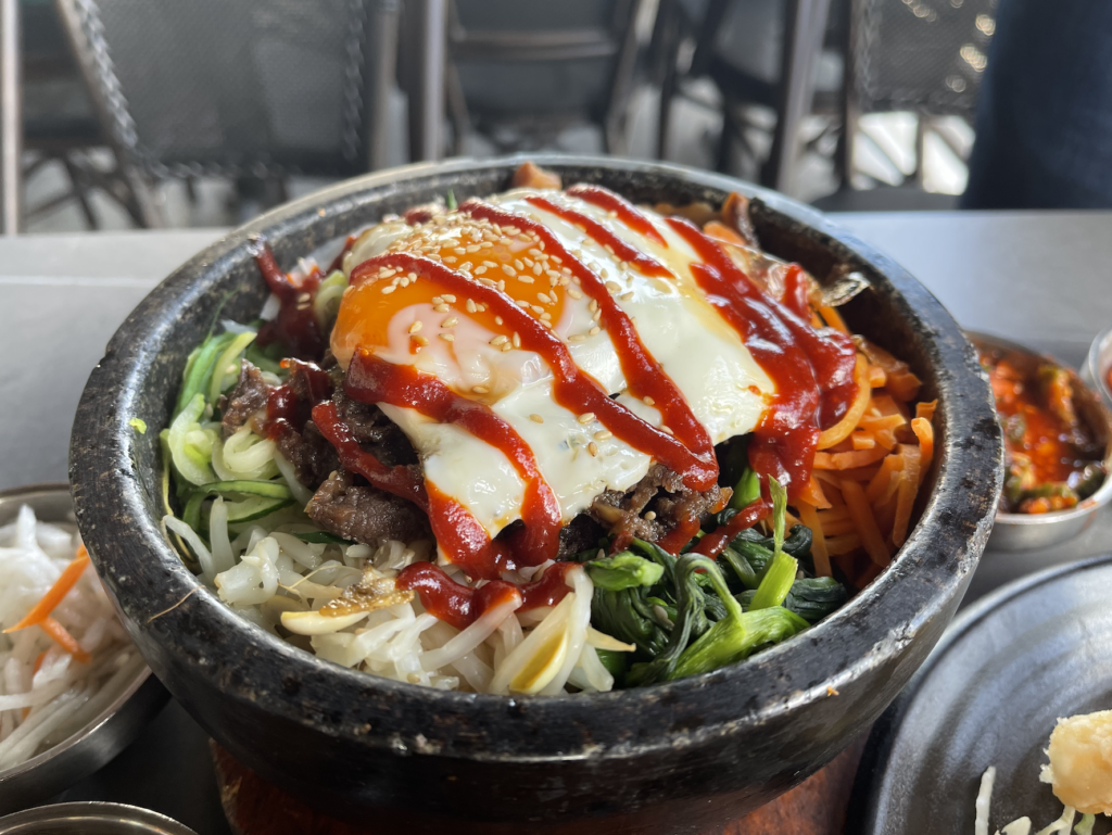 Beef bulgogi bibimbap is served in a stone bowl and layered with rice, wilted spinach, matchstick carrots, cooked bean sprouts then topped with the marinated diced meat and a fried egg. Spicy gochujang paste is stripped over top.