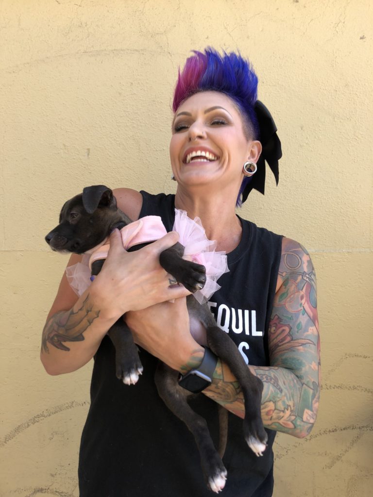 woman with magenta-and-blue hair, in black tank top, holds little black dog. Woman's arm is tattooed.