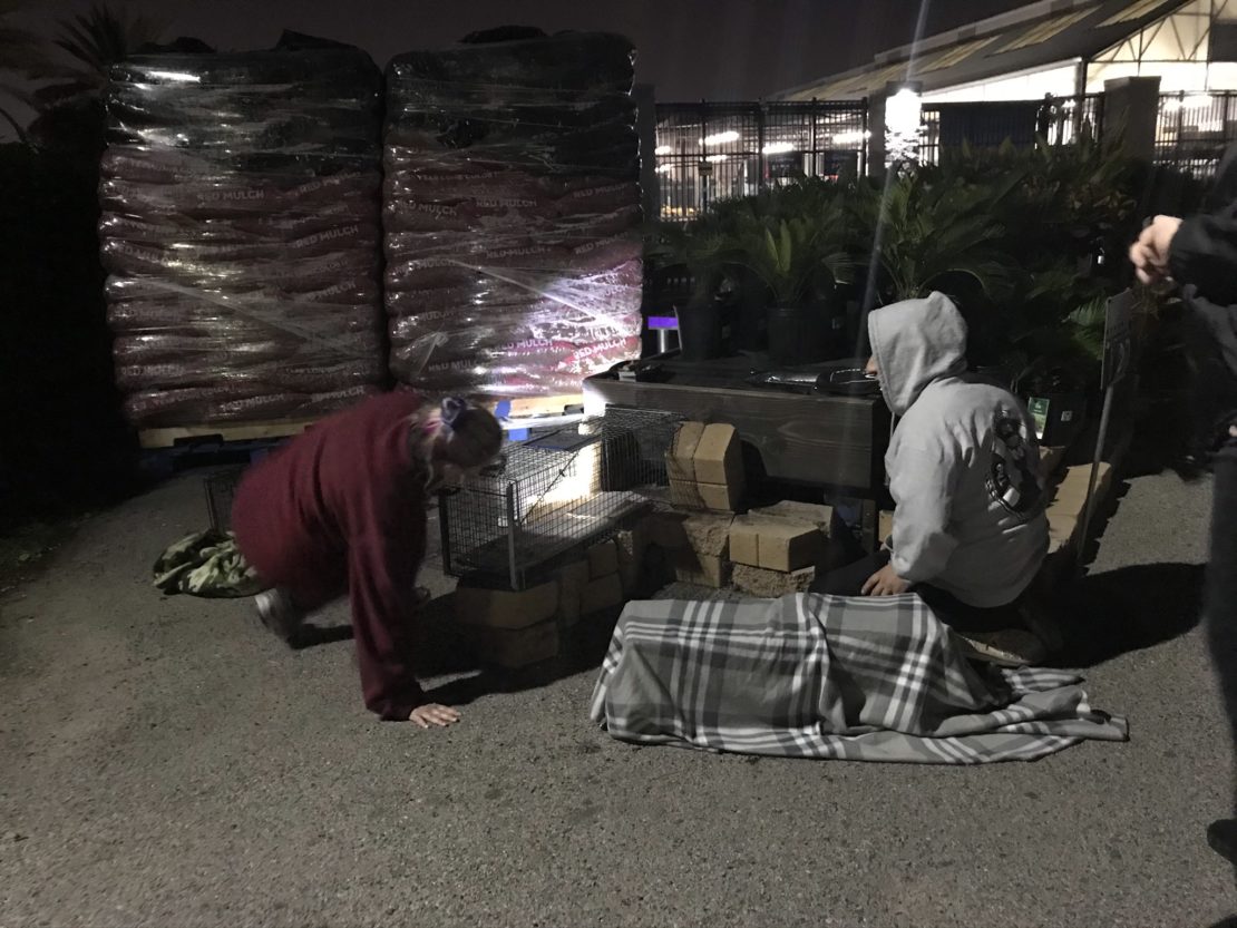 people in hoodies and sweatshirts at night, readying a trap to catch a kitten. Several bags of fertilizer in background.