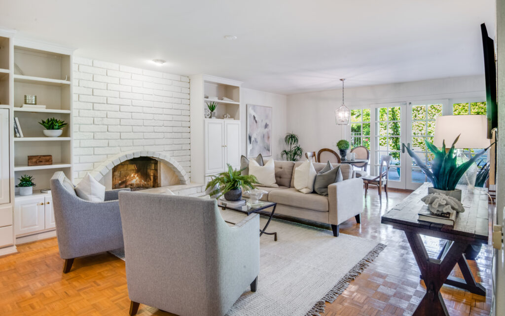 A bright id-Modern living room with parquet flooring. Gray chairs and a sofa are arranged around a glass coffee table over a light blue rug. A white brick fireplace is brackets by floor-to-ceiling white shelving holding plants and artwork.