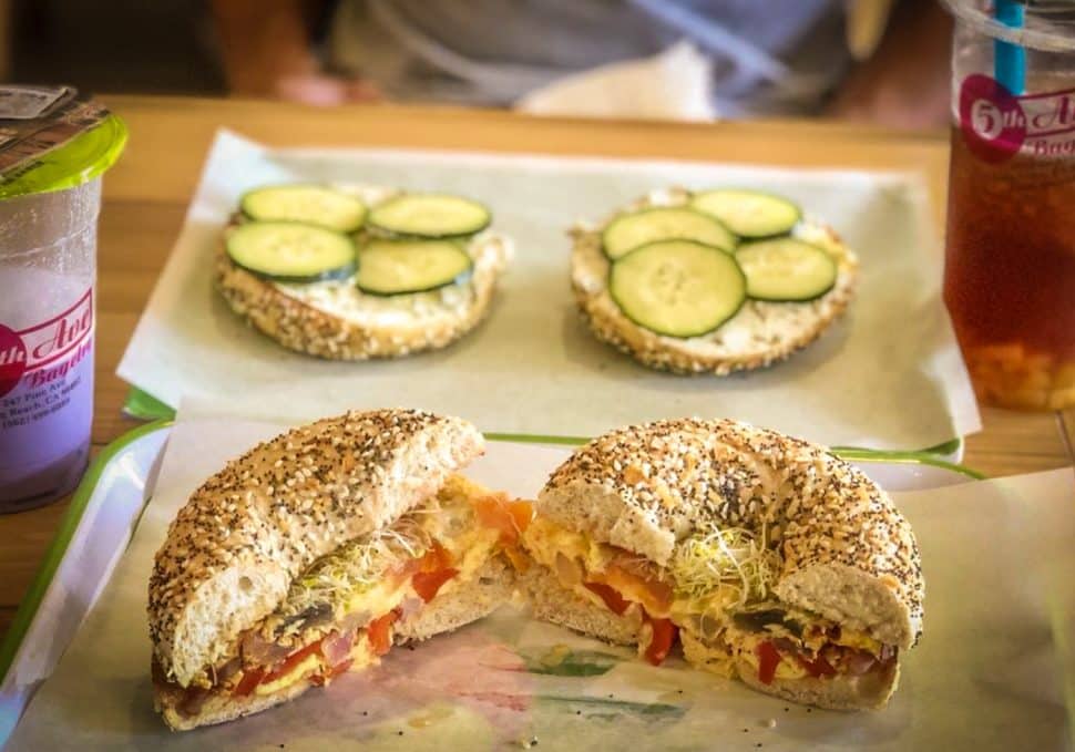 Two plates with bagels; one as a breakfast sandwich and the other covered with cream cheese and cucumbers. Two drinks flank each side.