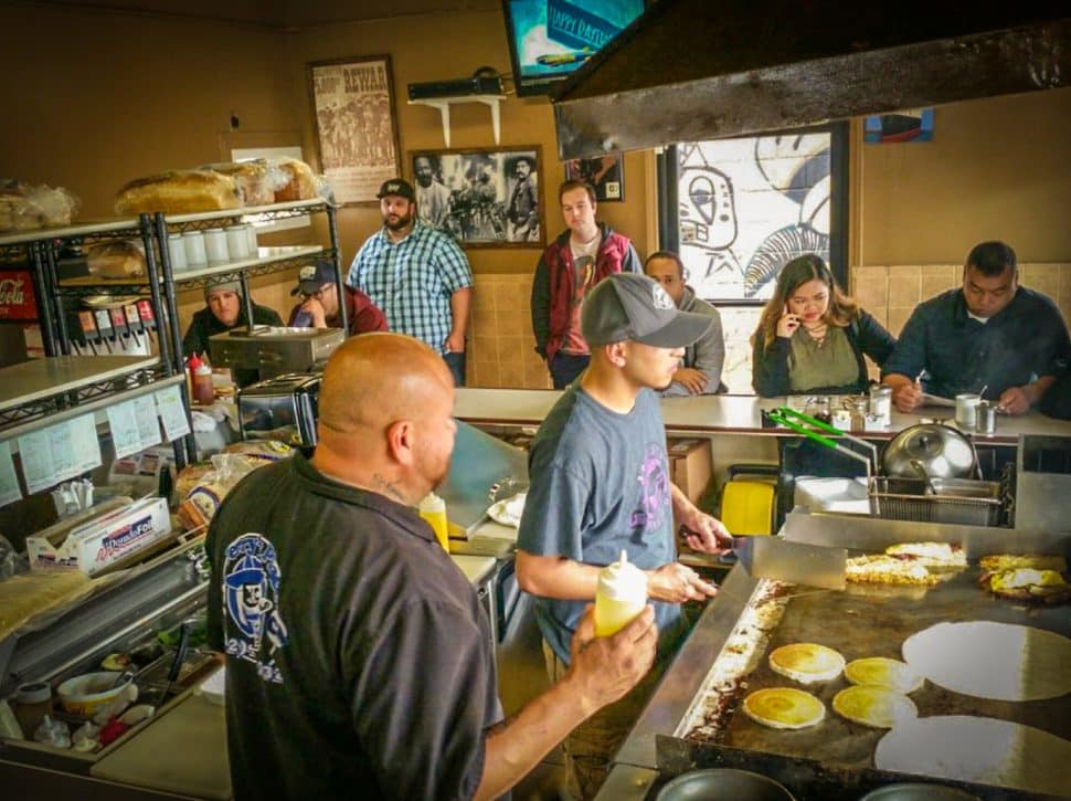 Two cooks stand a griddle inside a small restaurant space surrounded by customers.