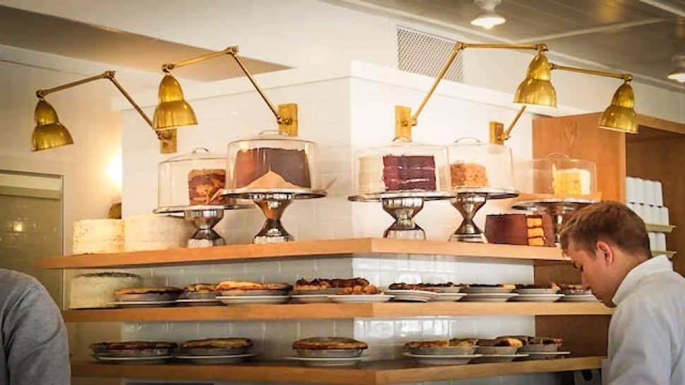 White interior of a bakery and restaurant has open-air shelves lined with various cakes and pies.