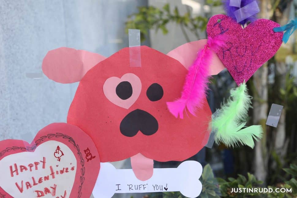 Valentine card made of heart shapes and shaped into a dog with pink ears, an upside-down red heart face, two black eyes with a pink heart-shaped patch over one, a black upside down heart nose, a pink tongue, and a white bone with "I love you" written in ink.