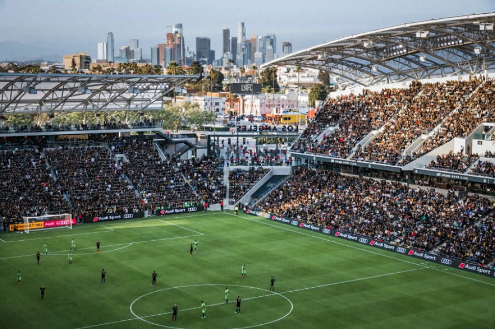 The buildings of Downtown LA act as a backdrop to a giant stadium filled with people watching a soccer game.