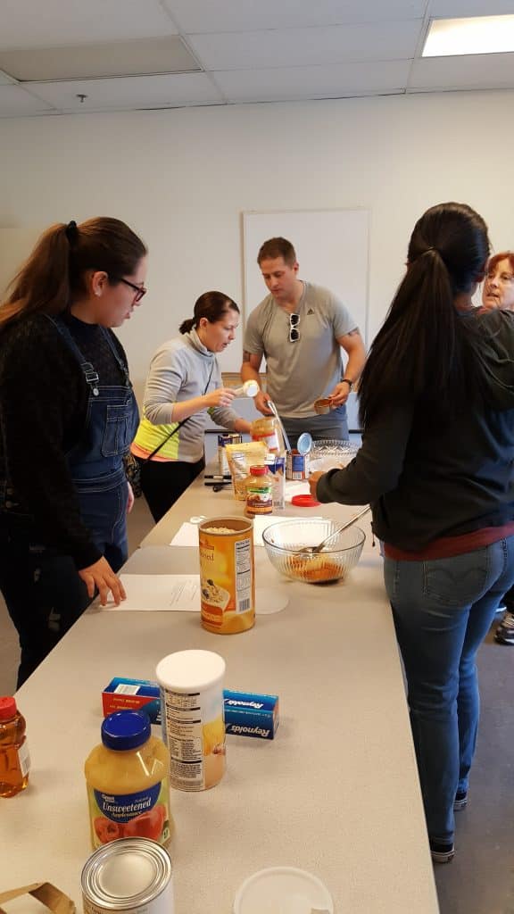 long table with a woman with a ponytial on one side, two volunteers at the end--a woman and a man, and a fourth in front. Jars of peanut butter and dog treats on table. They are making treats for shelter dogs.