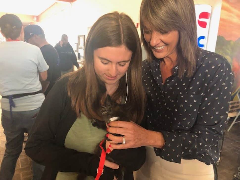 teenage girl with long brown hair with adult woman with long brown hair holding black kitten with red harness, whose head is nuzzled in.