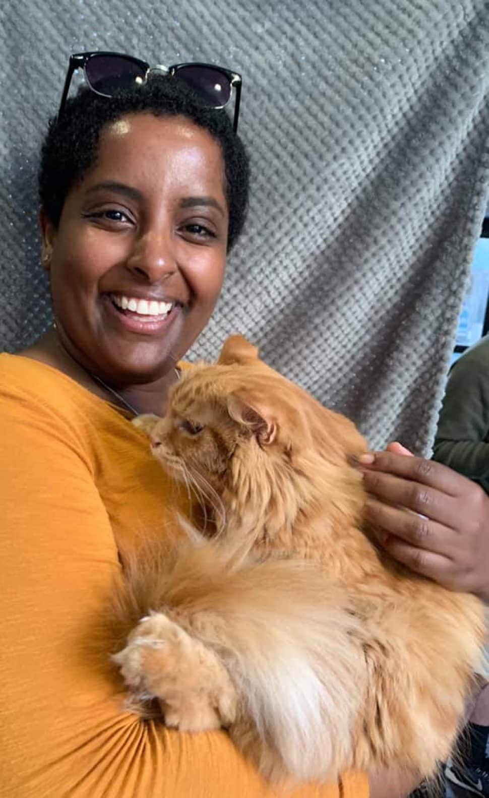 Woman with short black hair and sunglasses on top of her head, wearing a bright-orange shirt, cuddles newly adopted orange cat.